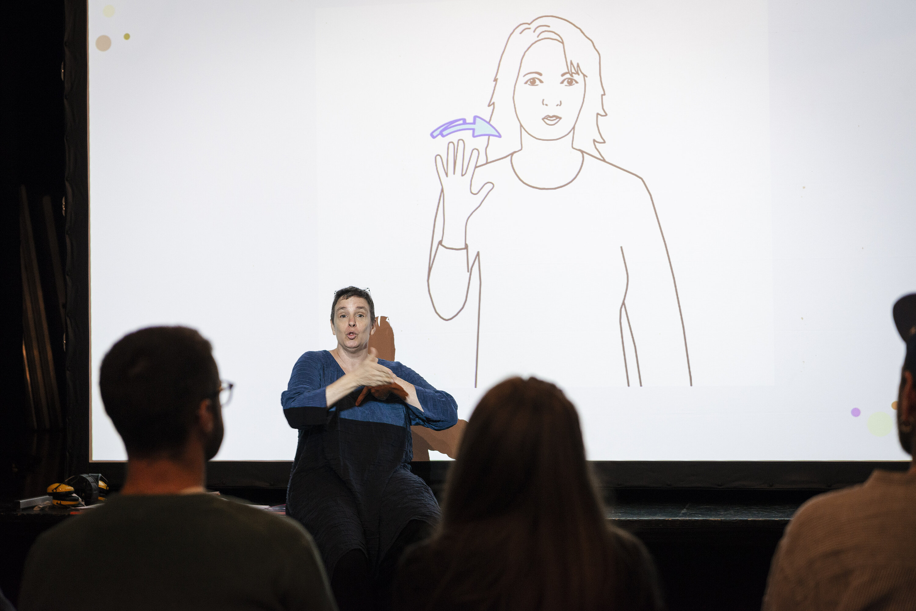 Lors de la sensibilisation à l’attention des institutions culturelles yverdonnoises pour les arts vivants, Anne-Claude Prélaz Giroud (co-directrice de l’Association Ecoute Voir) donne un atelier sur le handicap auditif. Sur l’image, elle fait une démonstration de la langue des signes. Derrière elle, un écran montre un dessin d’une personne qui fait le signe « d’au revoir ». ©Yverdon-les-Bains/ Pauline Stauffer, 2023 