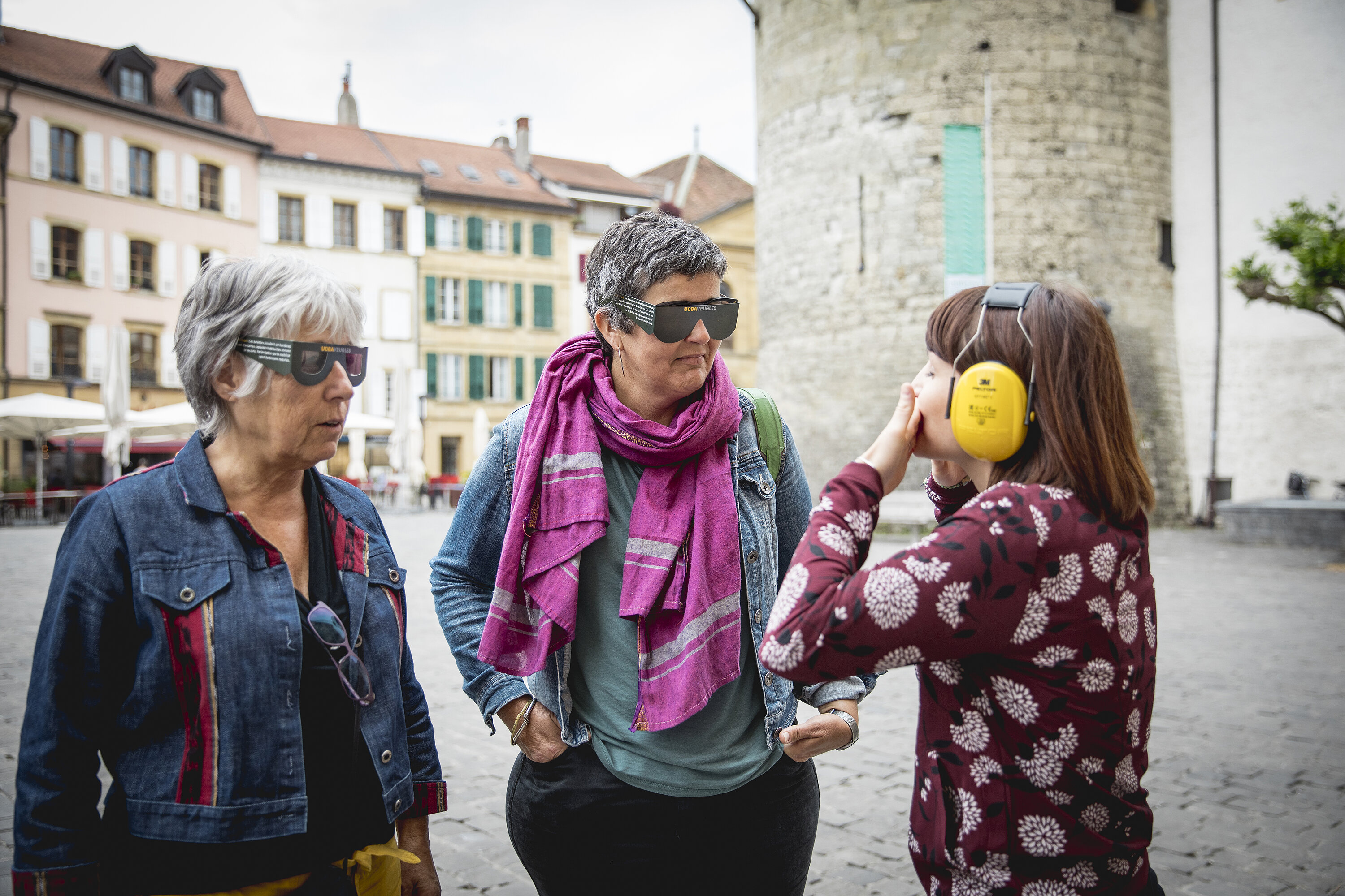 Un groupe de trois personnes se trouve sur la place Pestalozzi participer au projet pilote Yverdon culture accessible. Dans le cadre de la formation à la chaîne de l’accessibilité, la personne de gauche porte des lunettes pour simuler une déficience visuelle grave et la personne du milieu a des lunettes de vision tubulaire. La personne de droite a posé ses deux mains sur sa bouche et porte un casque de protection auditive. ©Yverdon-les-Bains/ Zoé Jobin, 2022 